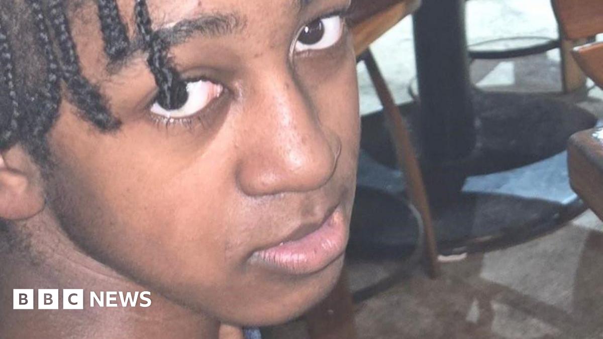 A snapshot of a young man with braided hair, looking into the camera from below. He appears to be in a bar or cafe, with the bases of tables visible in the background.