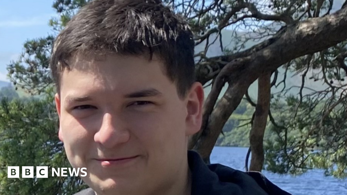 Tom Dingle smiling for a photograph. He has short dark brown/ black hair and is standing in front of a tree and a body of water.