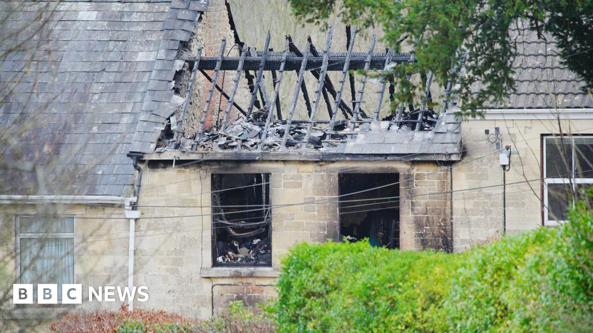 The charred shell of the destroyed home following a fire. The roof has collapsed and only the burnt wooden beams are visible. The glass windows have been smashed and are completely blackened from smoke. The surrounding homes on the terrace are untouched.