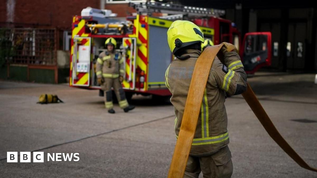 Firefighter officers running through a practice drill during a London Fire Brigade facility. One faces away from the camera holding a hose and another stands near a fire engine in the distance.