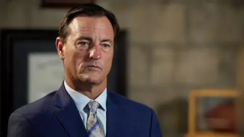 Brad Edwards, a man with short brown hair and a determined look on his face, pictured in a close-up portrait with the background blurred. He wears a navy suit, a pale blue shirt and a blue and grey tie.