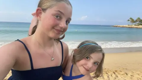 Family A selfie shows Ellie (left) on a beach with her younger sister Emilia (right). They are wearing summer dresses on a sunny day and the sand and sea are behind them.