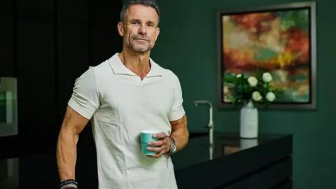 Toby Phillips Photography A man with greying hair in a white polo shirt. He is wearing a watch and some bracelets. He is holding a aqua-coloured cup and leaning on a kitchen counter.  