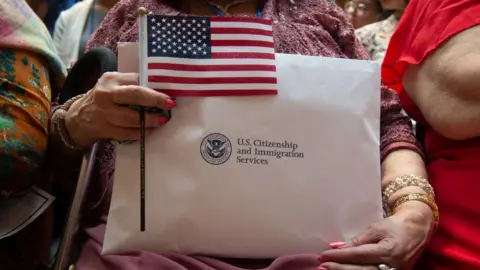 AFP via Getty Images A woman holding her citizenship papers from US Citizenship and Immigration Services, along with a small American flag, while waiting to take her oath
