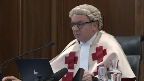 Judge Lord Colbeck, wearing a legal wig and uniform, sits in front of a laptop