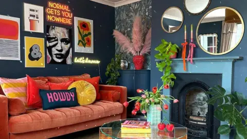 Rachel Verney A living room has a pink sofa with colourful cushions and multiple gold mirrors on the walls. 
