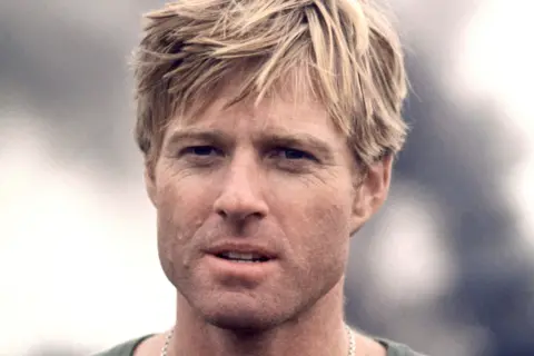 Getty Images Robert Redford, with short, tousled blonde hair is shown from the shoulders up, wearing a casual green top and a thin chain necklace. The background is softly blurred with muted gray tones, suggesting an outdoor setting.
