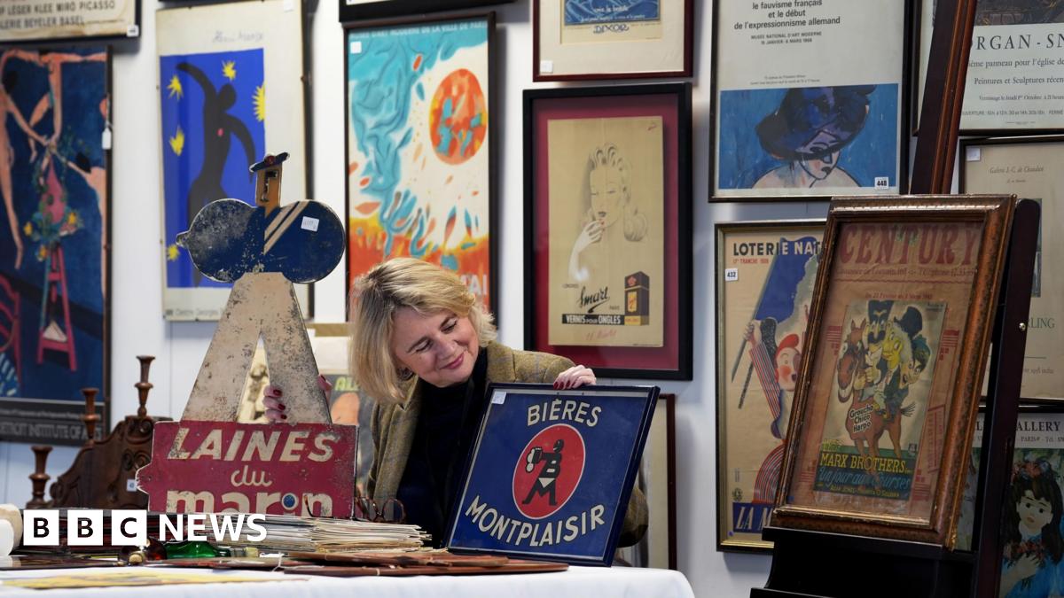 A woman sits at a table holding and looking at a enamel sign. All around her are prints and signs from the 20th century.