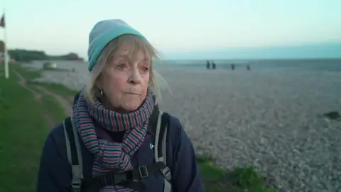A woman stood on a grassy bank in front of the pebble beach at Budleigh Salterton. She is wearing a blue hat, a striped scarf and a rucksack.