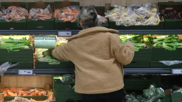 A woman with her back to the camera reaches into shelves of vegetables in a supermarket