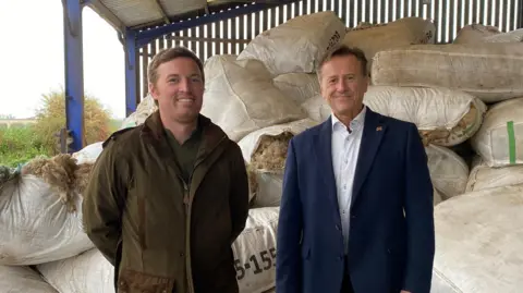 Staffordshire County Council Two men - one wearing a wax coat and the other wearing a suit with open collar - are standing in a barn with bags of wool stacked up behind them. They are both looking towards the camera and smiling.