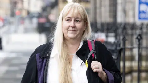 PA Media Maria Kelly has long blonde hair and is smiling. She is wearing a white shirt, dark jacket, and has a rucksack over her shoulder.