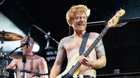 Getty Images James Johnston, of the band Biffo Clyro, performs on stage topless with pink trousers. His twin brother Ben, also topless, plays at a drum kit in the background.