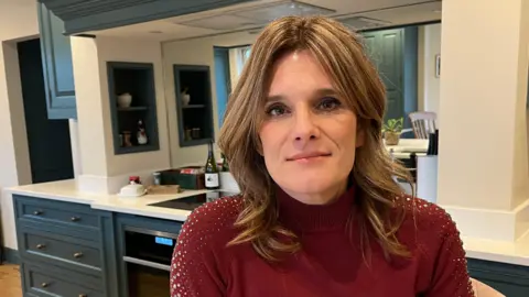 Ellen Roome in a burgundy top looking at the camera. She is in a kitchen and the counters and a stove are behind her.