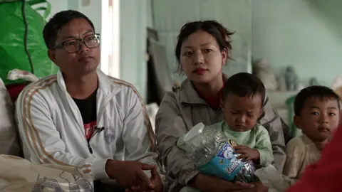 Bawi Nei Lian and his family sit on the floor of the community centre in India. He's dressed in a white track suit