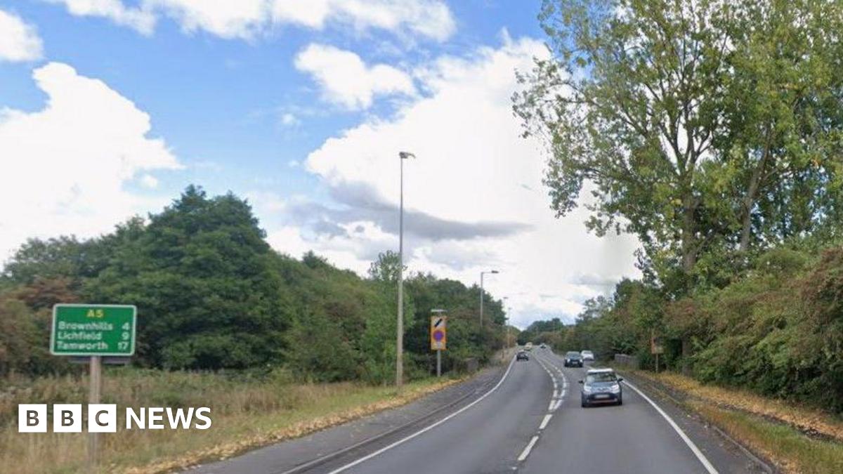 A road with vehicles travelling in both directions. There are street lights and green road signs at the side of the road as well as a pavement running for some of its length.