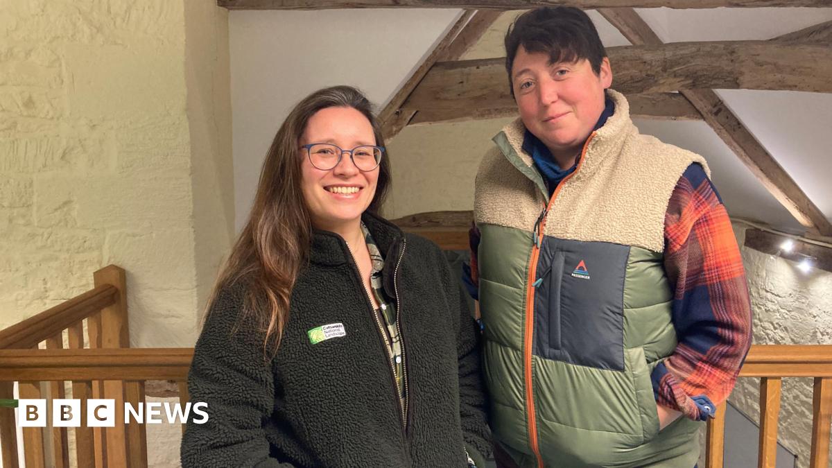 A woman with long brown hair over one shoulder, a black fleece with a white and green logo on it and blue glasses, stands beside a woman with short brown hair wearing a mostly green and cream coloured gilet with a blue and orange checked shirt beneath. They are both smiling and stood on a mezzanine level inside a converted barn.
