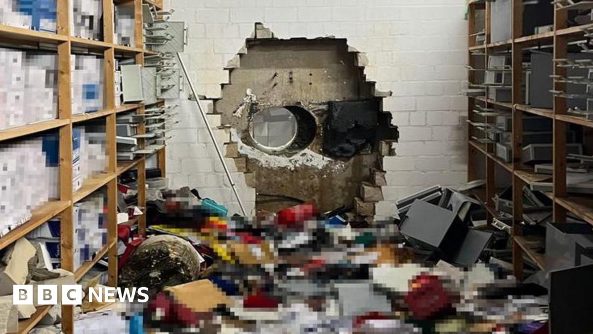 A large hole is seen in a white brick wall in the background, while many boxes are seen scattered on the floor around shelves in the foreground