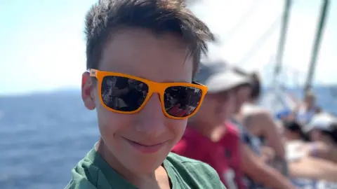 BBC Murray looking at the camera and smiling while wearing bright orange sunglasses. He appears to be on a boat, with the sea visible behind him and other people sitting alongside him.