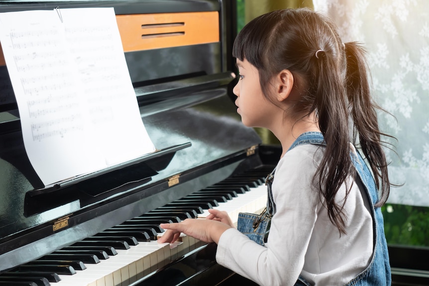 A young girl in pigtails and denim overalls sits at a piano with sheet music. Her hands are on the keys as she reads the music.