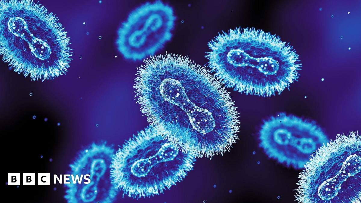 A representation of the mpox (monkeypox) virus under a microscope. Blue virus particles with fuzzy edges are swimming around against a dark blue background.