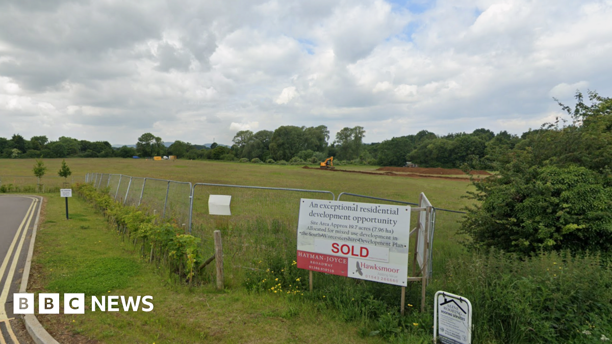 A white and red sign reads SOLD and is pitched against a metal fence. Behind the sign and fence is a large green field with one yellow digger moving piles of dirt.