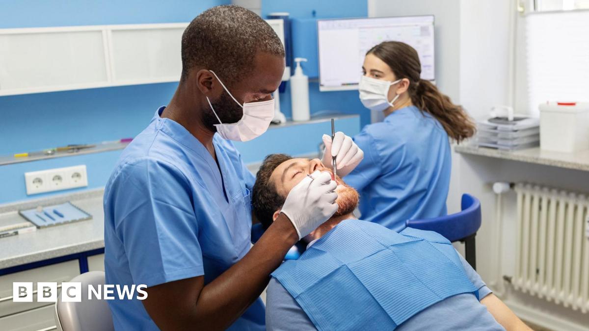 Male dentist examines the teeth of a male patient who is lying back the chair with his mouth open. A female assistant sits in the background.