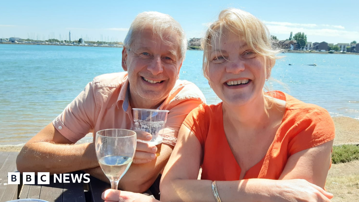 Chris and Ruth Stone-Houghton sit on a picnic bench, smiling into the camera. They are holding alcoholic drinks, and behind them is a body of water