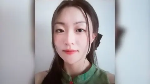 Met police picture of Zhe Wang wearing a green top. Her dark straight hair is tied back in a pony tail and she is look straight at the camera. 