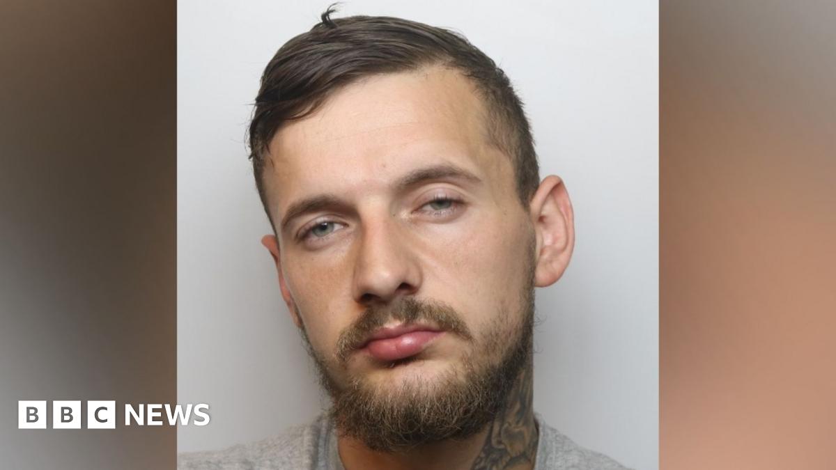 A mugshot of a man set on a blurred background. The man has short hair and a beard, and a neck tattoo is showing.
