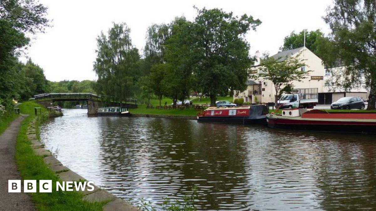 Canal with grassy path on left and red barges on right. Further right is a white pub with cars parked in front. A pedestrian bridge goes over the canal in the backdrop.