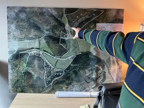 BBC/Mike Wendling A man, mostly outside the frame, points at a large map with sections parcelled out. Wooded areas and cleared areas are visible in shades of green.