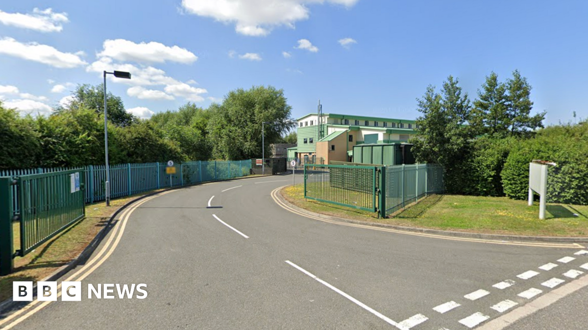 A road which enters into a gated site. Large buildings, which is used as the fire service's training centre, are in the distance.