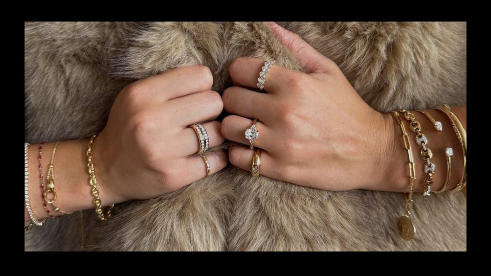 Various bracelets and rings on a pair of hands, in front of a light brown fur background.