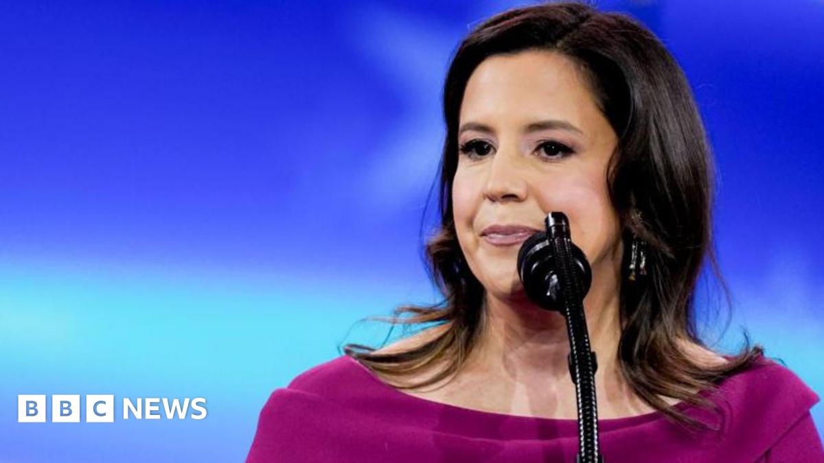 Representative Elise Stefanik stands before a microphone and in front of a blue backdrop. She wears a fuschia boat neck top.