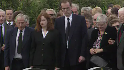 Cheryl Tooze and Jonathan Jones at the graveside. There are other mourners next to them. They are in black suits. 