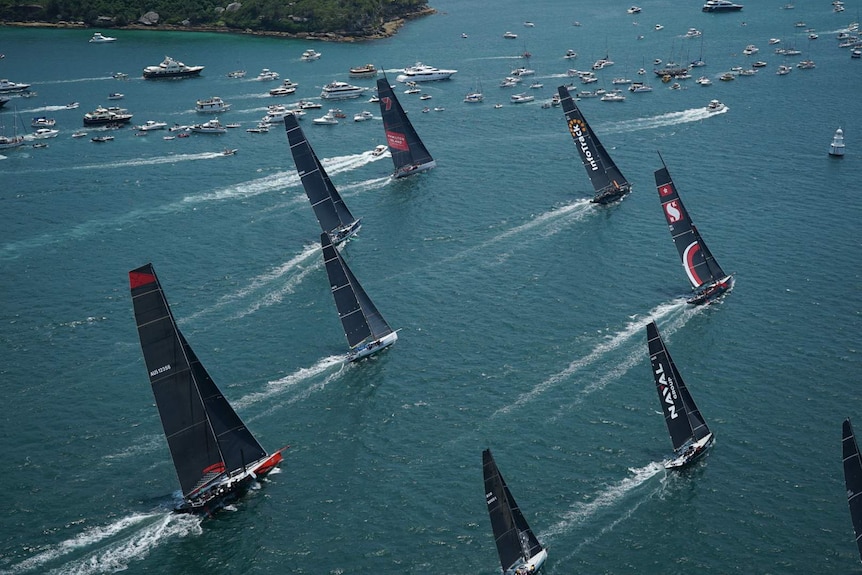 Yachts sail near land, with smaller boats in the background, as part of the 2019 Sydney to Hobart yacht race