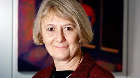 Anne Owers with medium-length brown hair looking at the camera with a slight smile on her face. She is wearing a red jacket over a black top. She is standing in front of a red picture hanging on a wall.