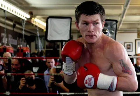 Getty Images Ricky Hatton is inside a boxing ring wearing bright red gloves and white hand wraps. The boxer is shirtless, showing a tattoo on the upper arm. The stance is forward-facing with fists raised in a defensive position. In the background, there are punching bags, photographers, and lighting equipment, indicating a training session or media event in a gym setting.