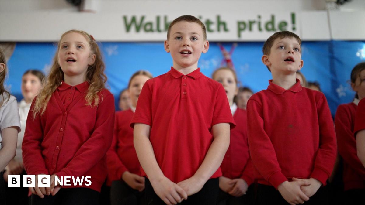 Students at Mersey Drive Community Primary School sing in their school hall. It's a close-up shot of three singing primary age children, two boys centre and right, and one girl on the left, all in red school uniform tops. More children stand behind them in rows in the choir.