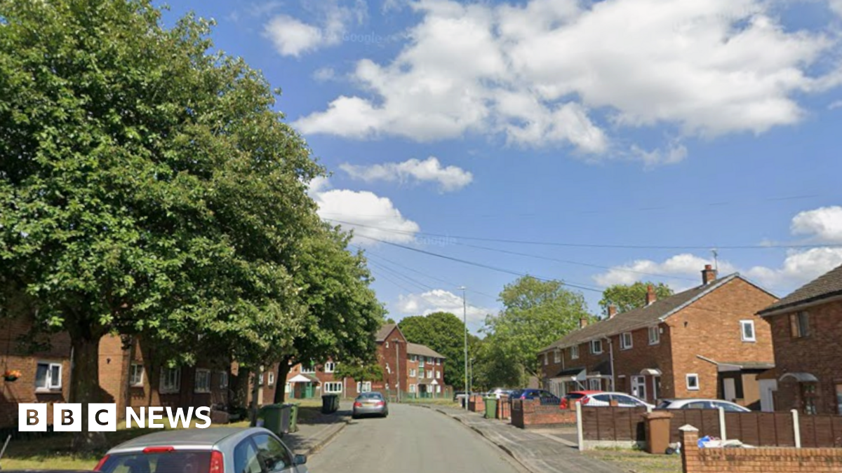 Large trees can be seen on the left of the street which has houses either side with cars parked in the road or on some drives. The sky is blue with some clouds.