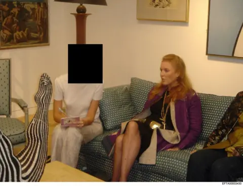 US Department of Justice Sarah Ferguson, wearing a purple coat and with her hair worn long, sits on a green sofa next to a woman who is wearing a short sleeved white t-shirt and writing in a notebook