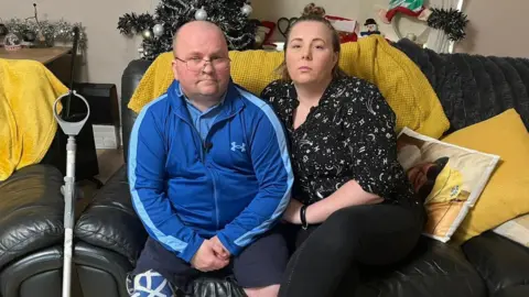 BBC An image of two people sitting on a sofa, with Christmas decorations in the background. On the left is a bald man wearing glasses and a tracksuit jacket; his right leg is a prosthetic decorated with Saltire flags and a crutch leans against the couch next to him. On the right, a woman leaning against him, wearing a patterned shirt, with her hair in a bun. 