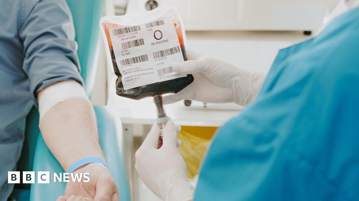 A person undergoing a blood transfusion in hospital. They are lying on a hospital bed, with their arm visible. A nurse is checking the blood next to them.