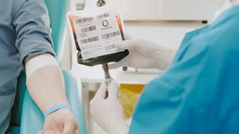 Getty Images A person undergoing a blood transfusion in hospital. They are lying on a hospital bed, with their arm visible. A nurse is checking the blood next to them.
