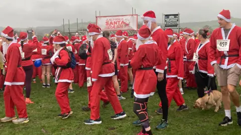 Hundreds of people dressed up in Santa outfits at the start line of the run. It is a very grey day, and people are holding up their phones to take pictures at the start line. 