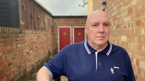 BBC A man with a shaved head, dressed in a blue polo shirt, stands in a narrow yard between two red-brick walls. Two red wooden doors with white frames can be seen in the background.