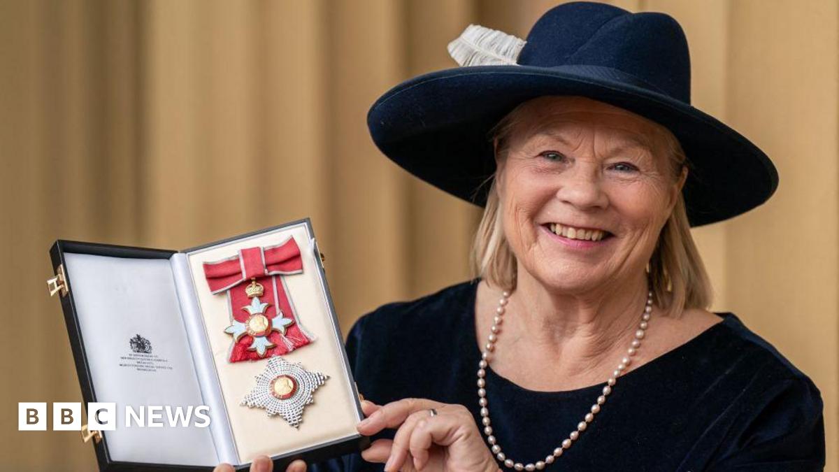 Dame Ann Limb, who wears a navy broad-rimmed hat with a white feather, has a blonde bob and wears a navy long-sleeved top with a pearl necklace, holds up her medal and smiles in a photo