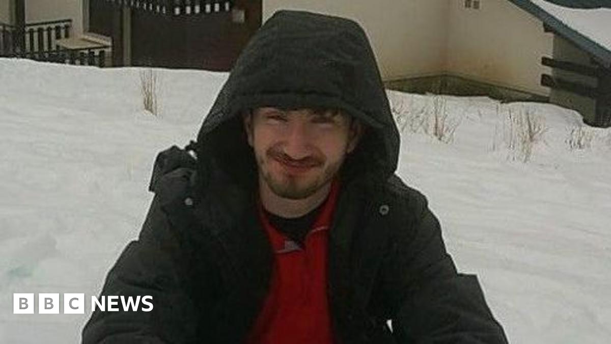 Matthew Pring poses for a photo in an outdoor area covered with deep snow. He is squatting and smiling and wears a dark anorak with the hood pulled over his head. He has brown hair and a short beard.
