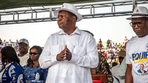 AFP via Getty Images Faustin-Archange Touadéra in a white shirt and hat stands in a stadium with his hands clasped. Supporters can be seen nearby.